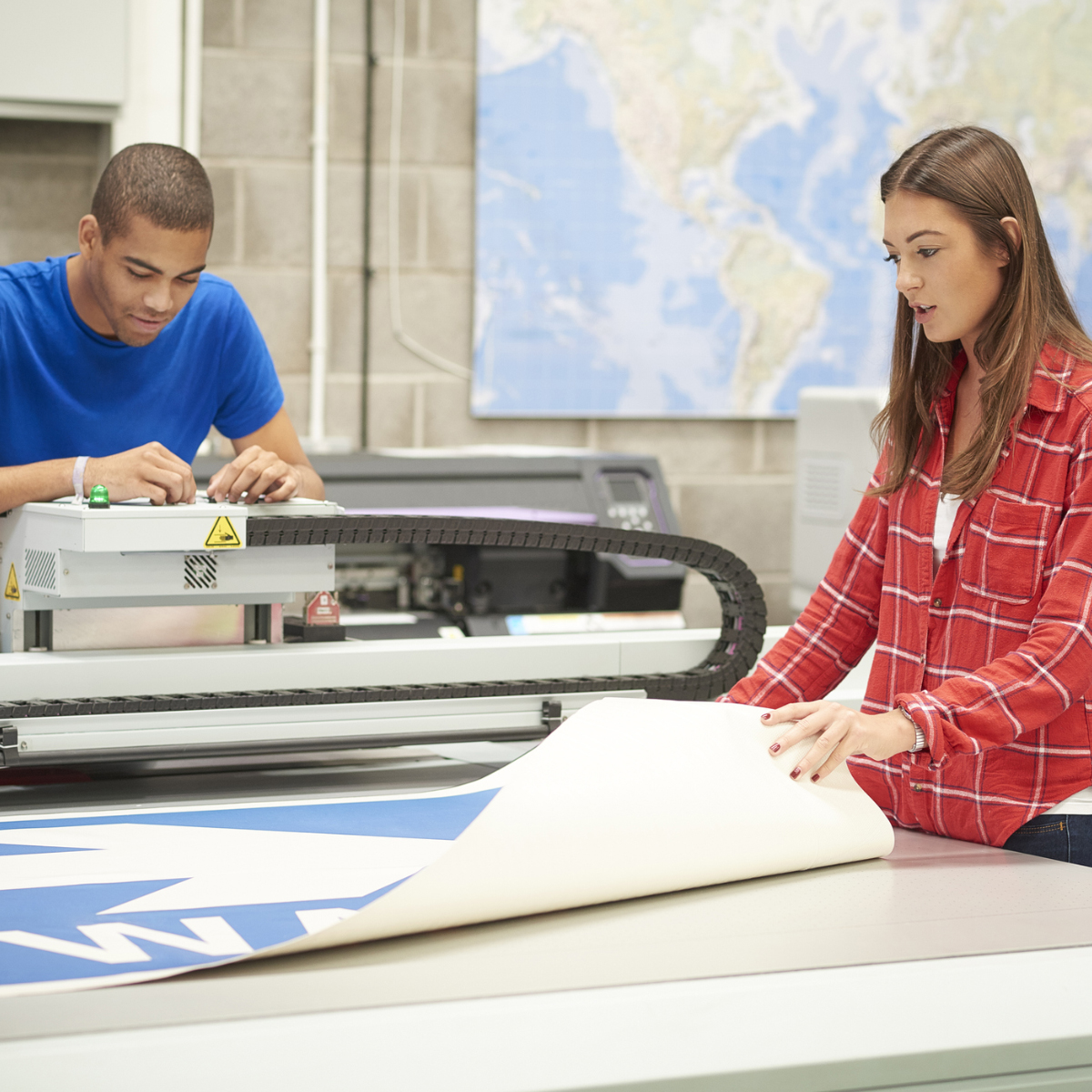 A young woman is working on a digital printing machine . she is printing out a large one way sign . A colleague is resetting the printer heads . ** background map has been modified**
